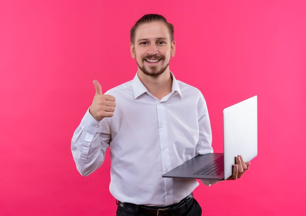 Apuesto hombre de negocios en camisa blanca sosteniendo el portátil mirando a la cámara sonriendo mostrando los pulgares para arriba sobre fondo rosa