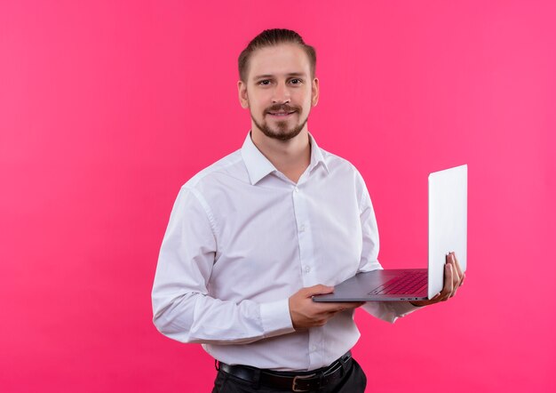 Apuesto hombre de negocios en camisa blanca sosteniendo portátil mirando a cámara con sonrisa de confianza de pie sobre fondo rosa