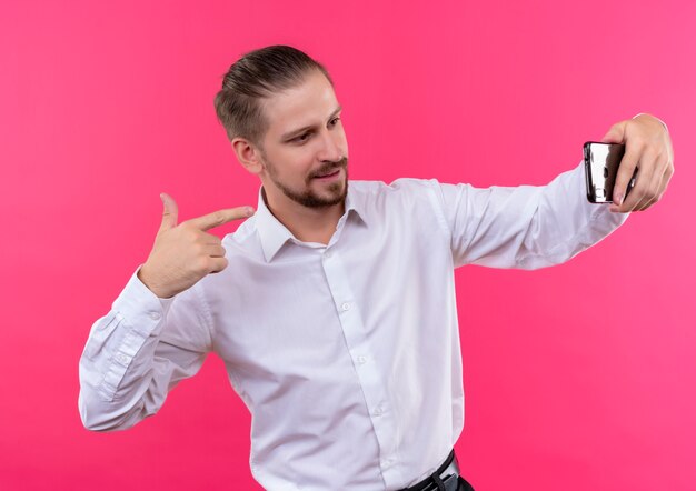 Apuesto hombre de negocios en camisa blanca tomando selfie con su smartphone sonriendo de pie sobre fondo rosa