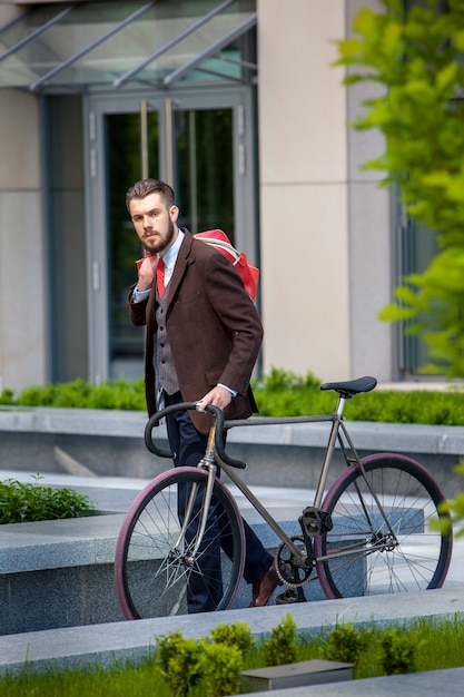 Apuesto hombre de negocios en una chaqueta y bolso rojo y su bicicleta en las calles de la ciudad.
