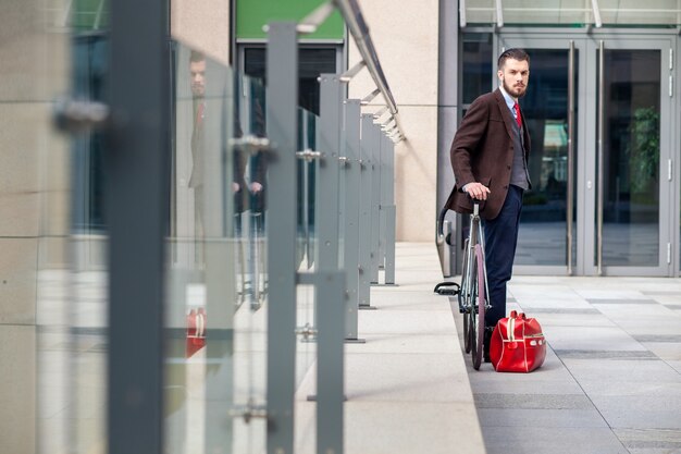 Apuesto hombre de negocios con chaqueta y corbata roja y su bicicleta en las calles de la ciudad. bolsa roja se encuentra a continuación. El concepto del estilo de vida moderno de los hombres jóvenes.
