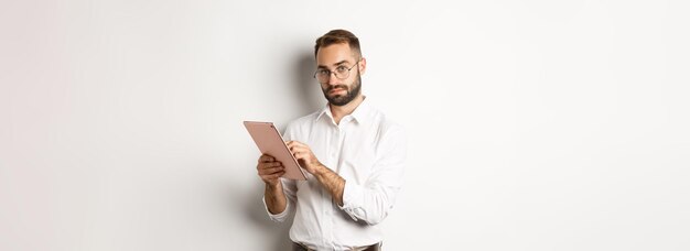 Apuesto hombre de negocios haciendo trabajo en tableta digital leyendo algo de pie sobre fondo blanco.