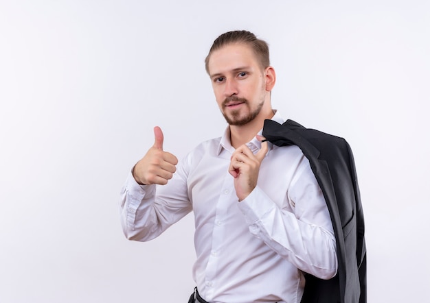 Foto gratuita apuesto hombre de negocios llevando su chaqueta sobre el hombro mirando a la cámara con una sonrisa de confianza mostrando los pulgares para arriba sobre fondo blanco.