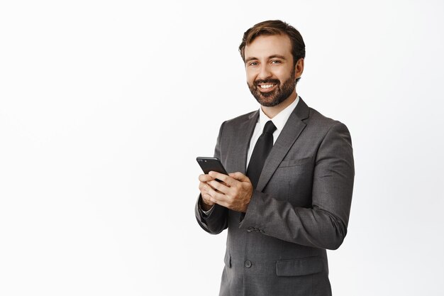 Un apuesto hombre de negocios sonriente usando un celular sosteniendo el teléfono en ambas manos y mirando complacido a la cámara de pie en traje sobre fondo blanco