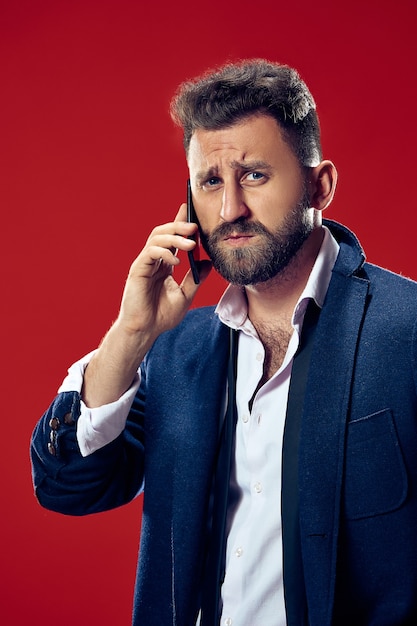 Apuesto hombre de negocios con teléfono móvil. Hombre de negocios serio que se encuentran aisladas en la pared roja. Hermoso retrato masculino de medio cuerpo. Las emociones humanas, el concepto de expresión facial.