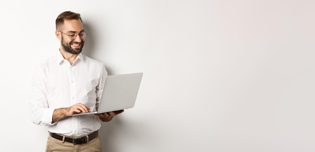 Apuesto hombre de negocios trabajando en una laptop respondiendo mensajes y sonriendo de pie sobre blanco b