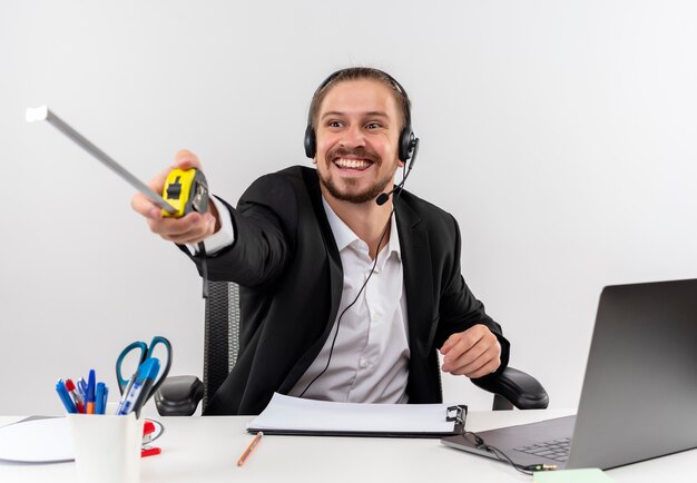 Apuesto hombre de negocios en traje y auriculares con un micrófono apuntando con cinta métrica al lado con una sonrisa en la cara sentado en la mesa en offise sobre fondo blanco.