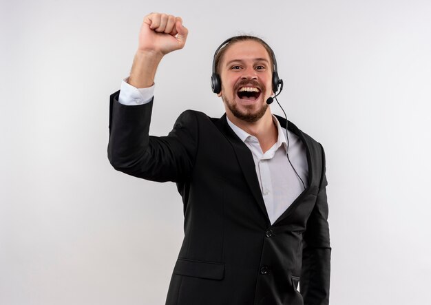 Apuesto hombre de negocios en traje y auriculares con un micrófono mirando a la cámara apretando el puño feliz y emocionado de pie sobre fondo blanco.