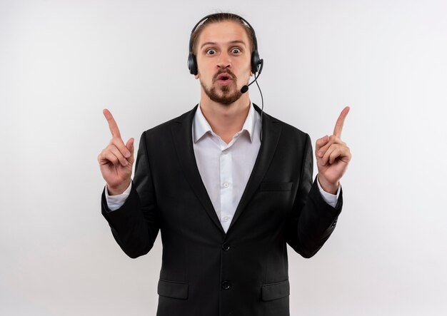 Apuesto hombre de negocios en traje y auriculares con un micrófono mirando a la cámara apuntando hacia arriba con los dedos índices sorprendido y feliz de pie sobre fondo blanco.