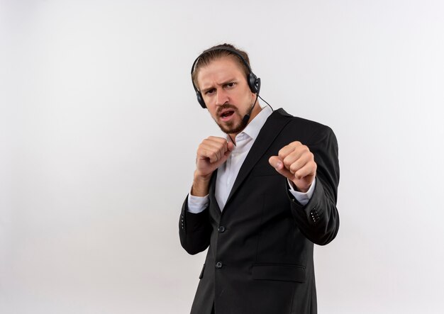 Apuesto hombre de negocios en traje y auriculares con un micrófono mirando a la cámara con cara seria posando como un boxeador de pie sobre fondo blanco.