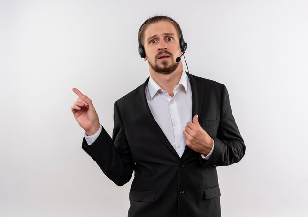 Apuesto hombre de negocios en traje y auriculares con un micrófono mirando a la cámara confundido apuntando con los dedos hacia el lado de pie sobre fondo blanco.