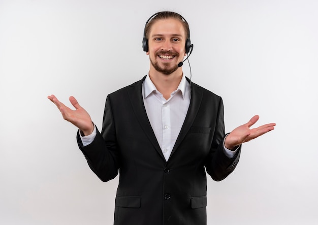 Apuesto hombre de negocios en traje y auriculares con micrófono mirando a cámara feliz y positivo sonriendo alegremente de pie sobre fondo blanco.