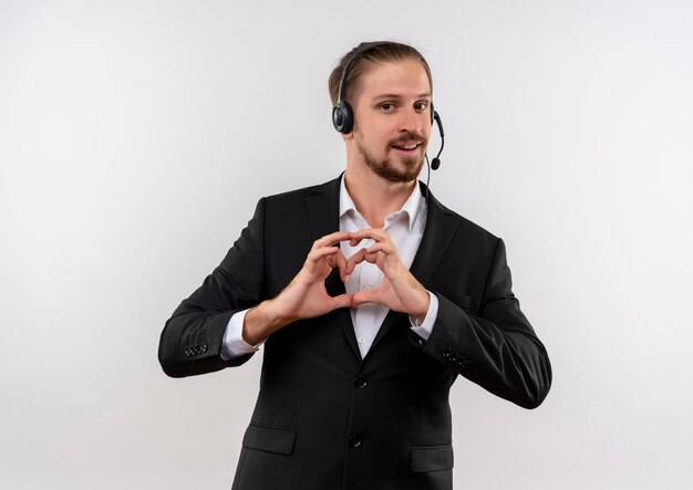 Apuesto hombre de negocios en traje y auriculares con un micrófono mirando a la cámara haciendo gestos herat con los dedos sobre el pecho sonriendo de pie sobre fondo blanco.