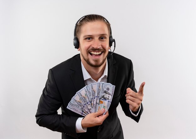 Apuesto hombre de negocios en traje y auriculares con un micrófono mirando a la cámara mostrando dinero en efectivo sonriendo alegremente de pie sobre fondo blanco.