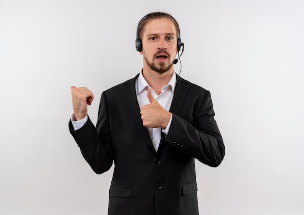 Apuesto hombre de negocios en traje y auriculares con un micrófono mirando a la cámara positiva y feliz apuntando con los dedos hacia el lado de pie sobre fondo blanco.