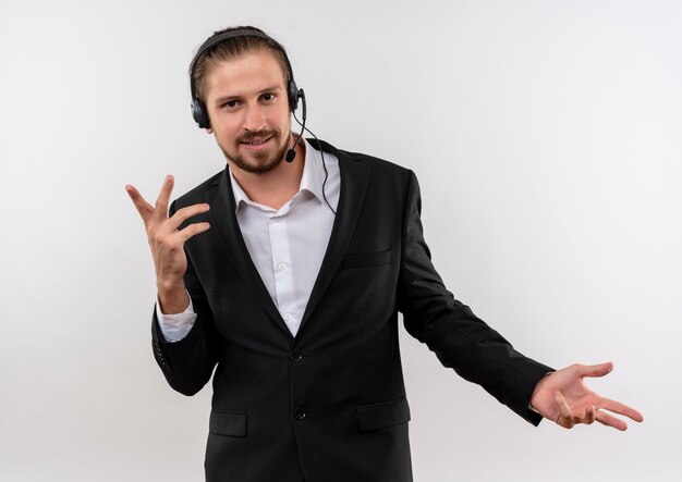 Apuesto hombre de negocios en traje y auriculares con un micrófono mirando a la cámara con una sonrisa en la cara gesticulando con las manos de pie sobre fondo blanco.