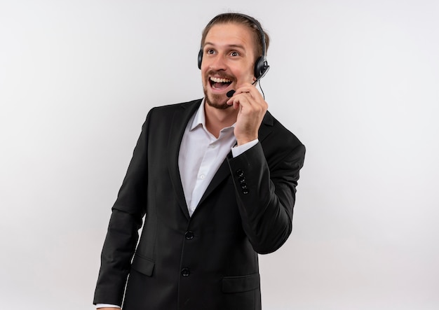 Apuesto hombre de negocios en traje y auriculares con un micrófono mirando a un lado escuchando a un cliente sonriendo con cara feliz de pie sobre fondo blanco.