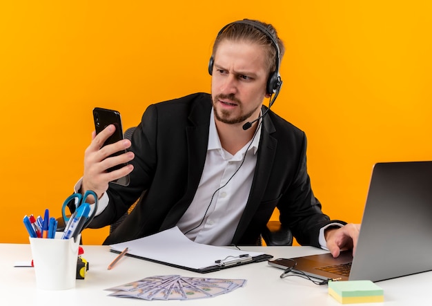 Apuesto hombre de negocios en traje y auriculares con un micrófono mirando la pantalla de su teléfono inteligente con rostro serio sentado en la mesa en offise sobre fondo naranja