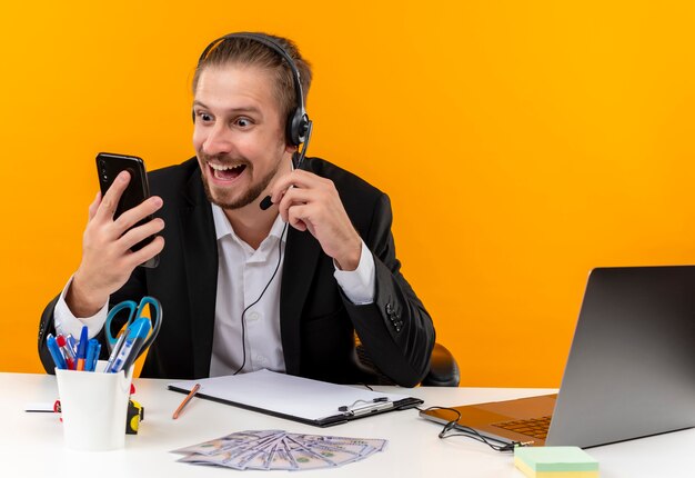 Apuesto hombre de negocios en traje y auriculares con un micrófono mirando la pantalla de su teléfono inteligente sorprendido y emocionado sentado en la mesa en offise sobre fondo naranja