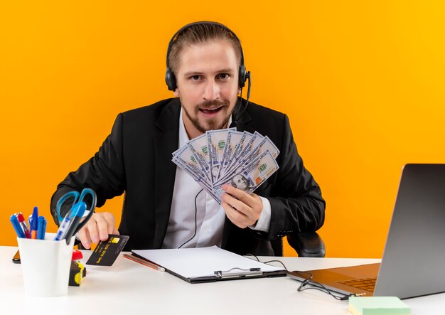 Apuesto hombre de negocios en traje y auriculares con un micrófono mostrando cojín mirando a la cámara feliz y emocionado sentado en la mesa en offise sobre fondo naranja