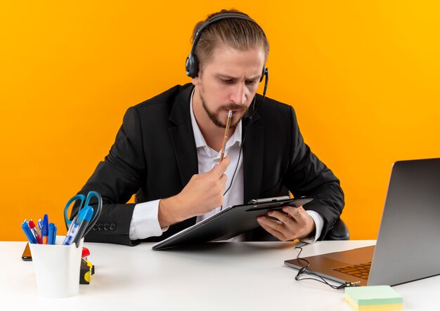 Apuesto hombre de negocios en traje y auriculares con un micrófono con portapapeles con documets mirando con cara seria sentado en la mesa en offise sobre fondo naranja