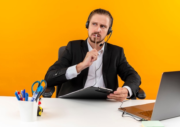 Apuesto hombre de negocios en traje y auriculares con un micrófono con portapapeles mirando a un lado desconcertado sentado en la mesa en offise sobre fondo naranja