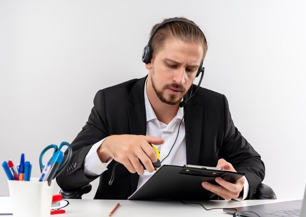 Apuesto hombre de negocios en traje y auriculares con un micrófono con portapapeles mirándolo con cara seria sentado en la mesa en offise sobre fondo blanco.