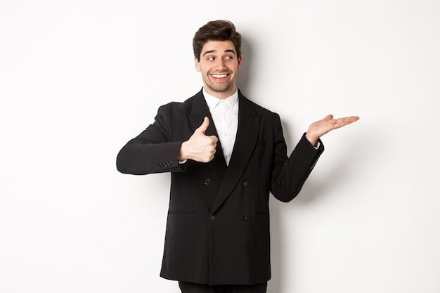 Apuesto hombre de negocios en traje negro, mostrando el pulgar hacia arriba y sosteniendo su producto en la mano sobre el espacio de la copia en blanco, de pie contra el fondo blanco.