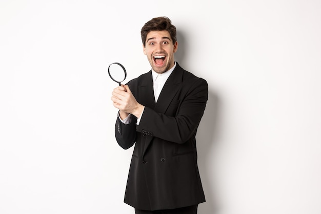 Apuesto hombre de negocios en traje negro, sosteniendo una lupa y sonriendo, encontró algo, de pie contra el fondo blanco.