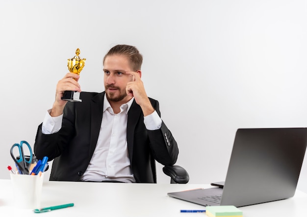 Apuesto hombre de negocios en traje sosteniendo el trofeo mirándolo feliz y complacido sentado en la mesa en offise sobre fondo blanco.