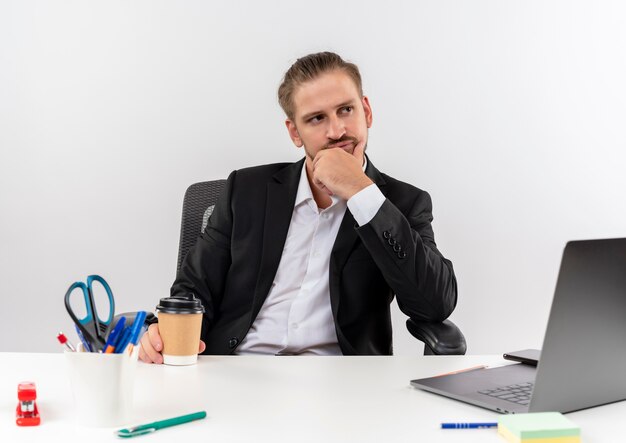 Apuesto hombre de negocios en traje con taza de café mirando a un lado perplejo sentado en la mesa en offise sobre fondo blanco.