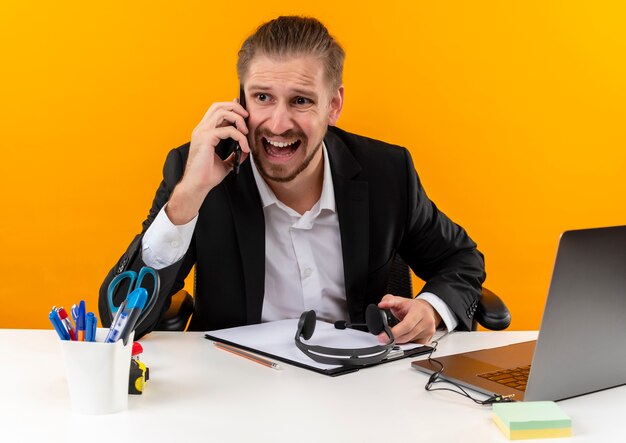 Apuesto hombre de negocios en traje trabajando en la computadora portátil mirando confundido y disgustado sentado en la mesa en offise sobre fondo naranja