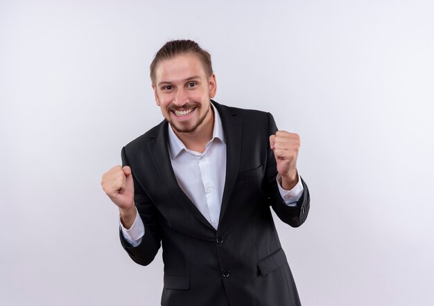 Apuesto hombre de negocios vestido con traje apretando los puños feliz y emocionado sonriendo alegremente de pie sobre fondo blanco.