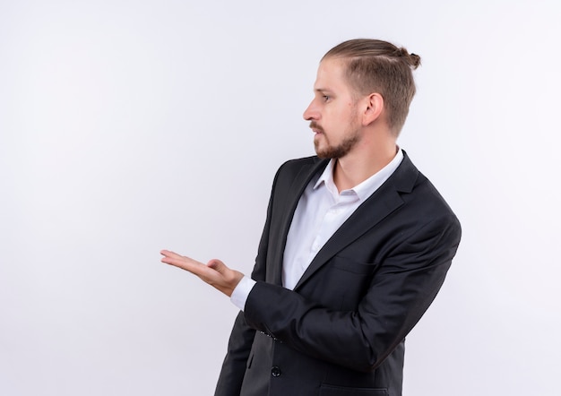 Apuesto hombre de negocios vestido con traje apuntando con el brazo de la mano hacia el lado de pie sobre fondo blanco.