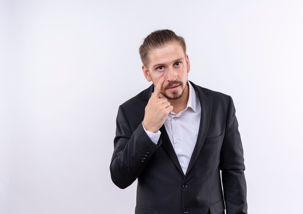 Apuesto hombre de negocios vestido con traje apuntando con el dedo a los ojos mirándote gesto de pie sobre fondo blanco.