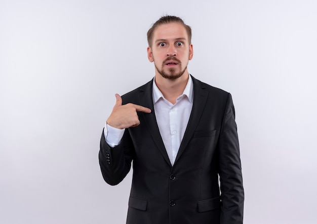 Apuesto hombre de negocios vestido con traje apuntando con el dedo a sí mismo mirando sorprendido de pie sobre fondo blanco.