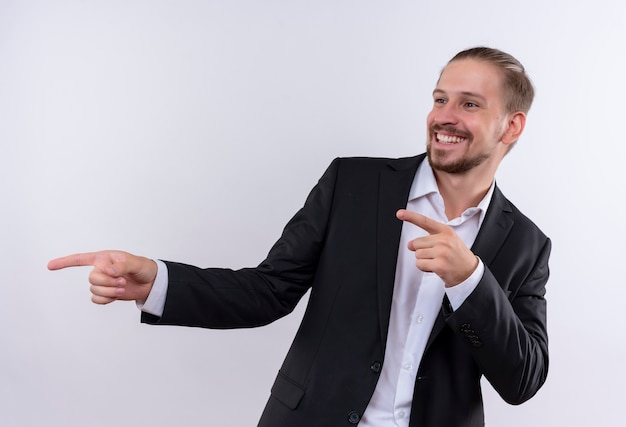 Apuesto hombre de negocios vestido con traje apuntando con los dedos hacia el lado feliz y positivo sonriente de pie sobre fondo blanco.