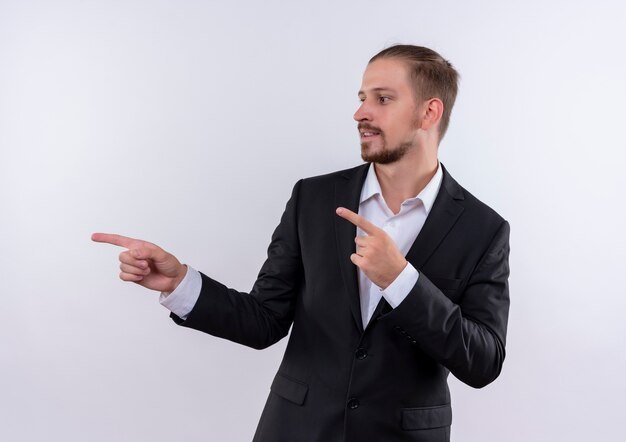 Apuesto hombre de negocios vestido con traje apuntando con los dedos hacia el lado mirando confiado de pie sobre fondo blanco.