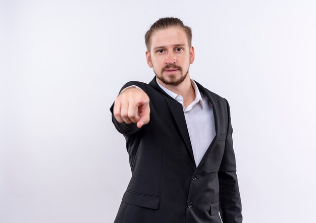 Apuesto hombre de negocios vestido con traje buscando confianza apuntando con el dedo índice a la cámara de pie sobre fondo blanco.