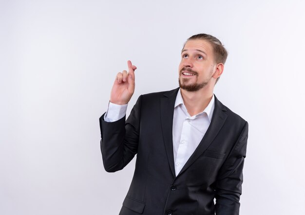 Apuesto hombre de negocios vestido con traje haciendo deseo deseable cruzar los dedos sonriendo de pie sobre fondo blanco.