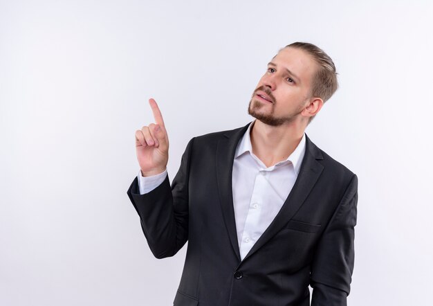 Apuesto hombre de negocios vestido con traje mirando a un lado con expresión pensativa apuntando con el dedo hacia arriba de pie sobre fondo blanco.