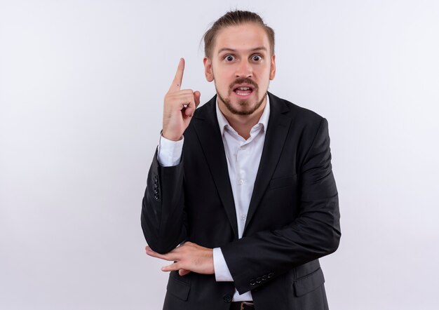 Apuesto hombre de negocios vestido con traje mostrando el dedo índice mirando sorprendido de tener una gran idea nueva de pie sobre fondo blanco.