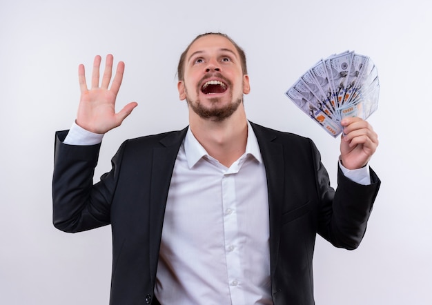 Apuesto hombre de negocios vestido con traje mostrando dinero en efectivo mirando hacia arriba feliz y emocionado de pie sobre fondo blanco.