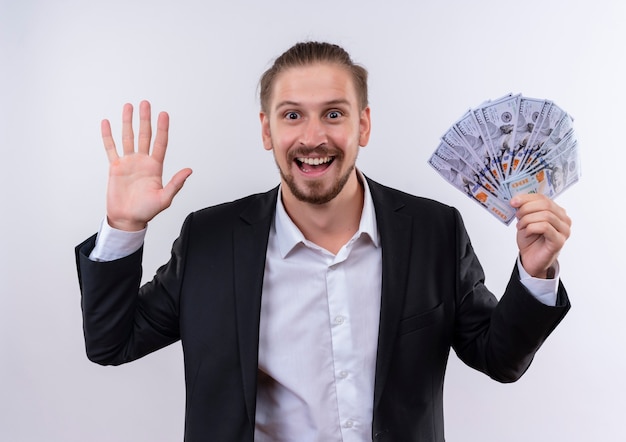 Apuesto hombre de negocios vestido con traje mostrando dinero en efectivo mirando a la cámara feliz y emocionado saludando con la mano de pie sobre fondo blanco.