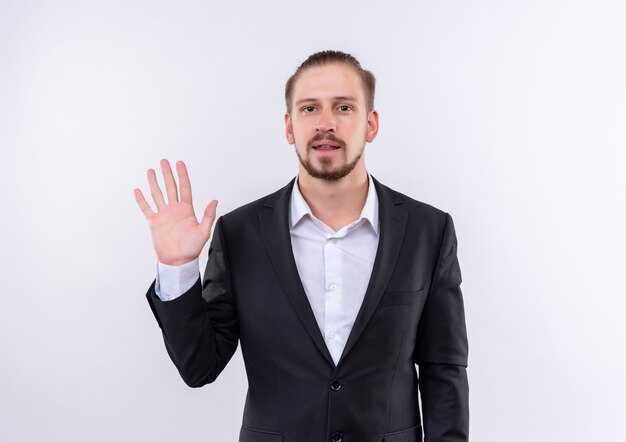 Apuesto hombre de negocios vestido con traje mostrando el número cinco sonriendo confiado de pie sobre fondo blanco