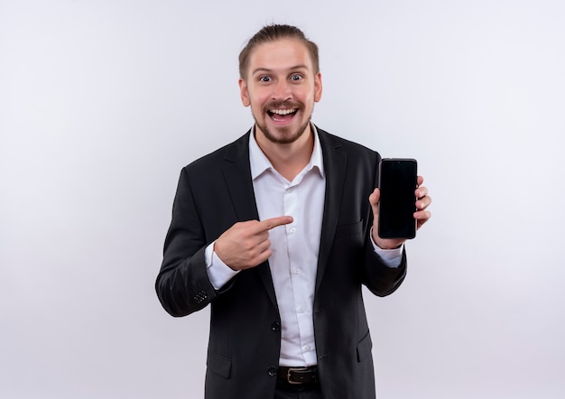 Apuesto hombre de negocios vestido con traje mostrando smartphone apuntando con el dedo sonriendo alegremente de pie sobre fondo blanco.