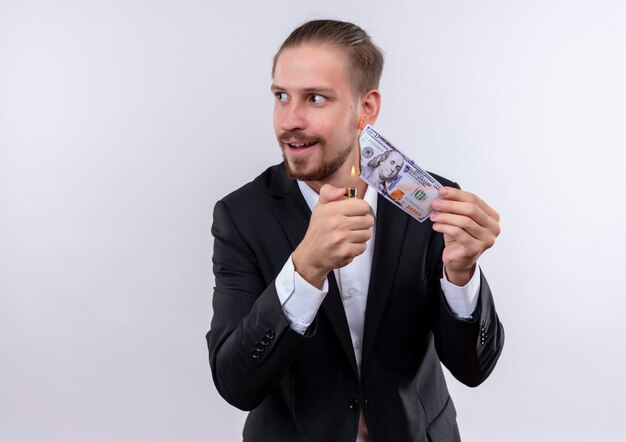 Apuesto hombre de negocios vestido con traje quemando dinero mirando a un lado con sonrisa astuta de pie sobre fondo blanco.