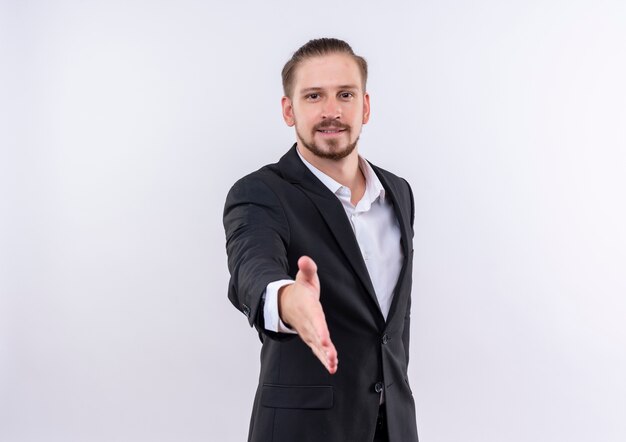 Apuesto hombre de negocios vestido con traje sonriendo amable saludo ofreciendo mano de pie sobre fondo blanco.