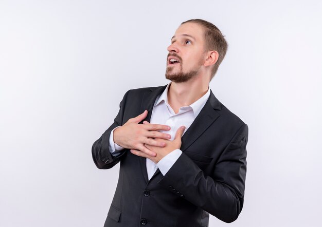 Apuesto hombre de negocios vestido con traje sosteniendo los brazos en el pecho sintiéndose agradecido de pie sobre fondo blanco.