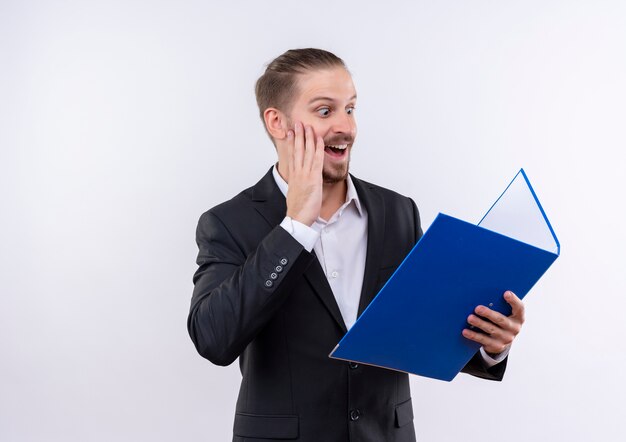 Apuesto hombre de negocios vestido con traje sosteniendo la carpeta mirando sorprendido y emocionado de pie sobre fondo blanco.